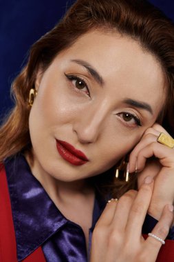 Kırmızı takım elbiseli ve ipek gömlekli şık bir Asyalı kadın kendine güvenen ve çekici bir ifadeyle poz veriyor, elleri zarif bir şekilde yüzünü çerçeveliyor..
