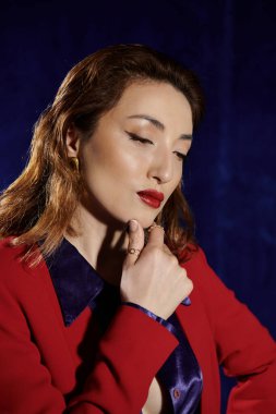 Uzun, dalgalı kahverengi saçlı, canlı kırmızı bir takım elbise ve ipek gömlekli bir kadın, koyu mavi bir arka plana doğru aşağı bakıyor..