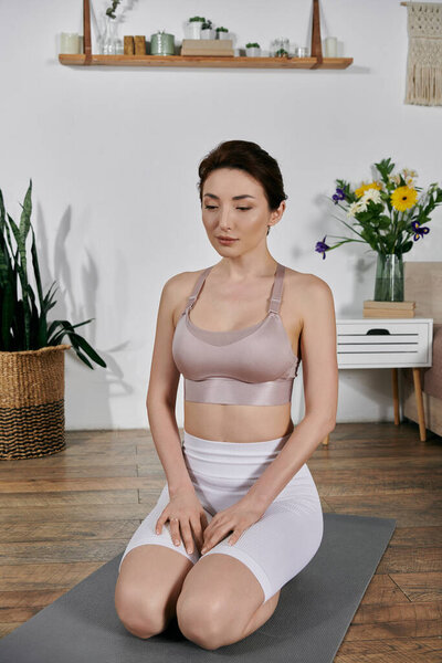 A beautiful Asian woman in a crop top meditates on a yoga mat in her modern apartment.