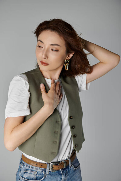 Уверенная молодая азиатка в зеленом жилете, белой футболке и джинсах позирует на сером фоне с распущенными волосами