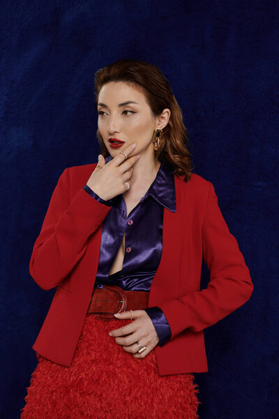 Стильная женщина позирует в красном костюме и шелковой рубашке на темном фоне, ее взгляд направлен в сторону.