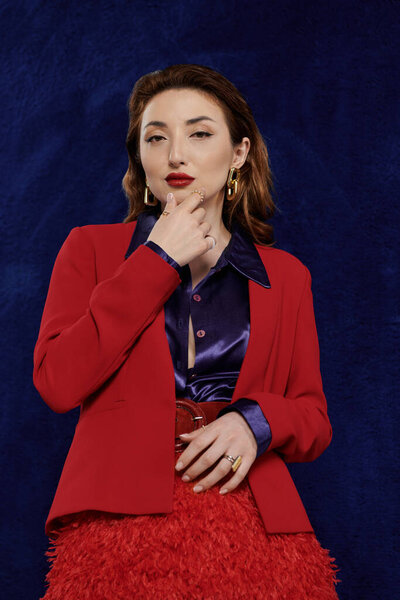 Привлекательная азиатка в ярком красном костюме и шелковой рубашке занимает уверенную позу на темно-синем фоне..