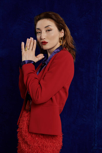 Стильная азиатка в ярком красном костюме стоит на тёмном фоне, её уверенный взгляд завораживает..