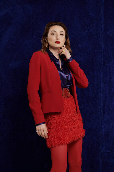 Азиатская женщина с уверенным взглядом позирует в ярко-красном костюме и текстурированной мини-юбке на темно-синем фоне.