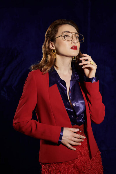 Стильная азиатка в красном костюме и шелковой рубашке позирует на тёмном фоне.