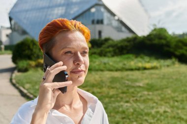 Turuncu saçlı bir kadın güneşli bir kampüste yürürken telefonla konuşuyor..