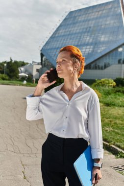 Kolej kampüsünde elinde mavi bir dosya olan beyaz tişörtlü bir kadın telefonla konuşuyor..