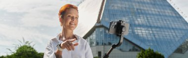 A woman with bright red hair smiles while filming with a selfie stick on a university campus. clipart