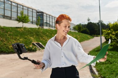 A teacher in a white shirt smiles while taking a selfie with a smartphone on a selfie stick. clipart