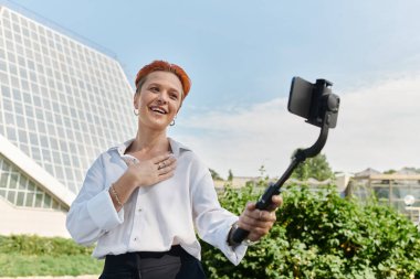 Beyaz gömlekli ve siyah pantolonlu bir öğretmen kampüste bir selfie videosu kaydederken gülümsüyor..