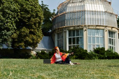 Renkli eşarplı bir kadın çimlerin üzerinde oturuyor, gülümsüyor..