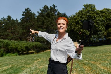 Kızıl saçlı bir kadın güneşli bir kampüste selfie çekerken kameraya gülümsüyor..