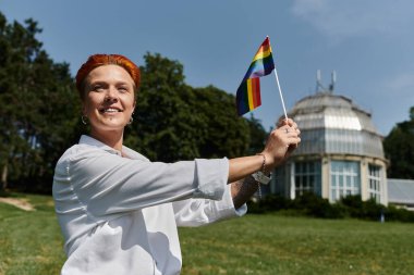 A woman smiles as she holds a rainbow flag in a park. clipart