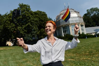 Gülümseyen bir kadın elinde gökkuşağı bayrağı ve kampüs binasının dışında selfie çekiyor..