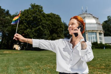 Gökkuşağı bayrağı taşıyan bir öğretmen kampüste telefonla konuşurken gülümsüyor..