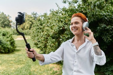 Beyaz gömlekli bir kadın dışarıda selfie çubuğuyla video çekerken gülümsüyor..