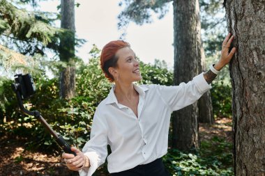 Beyaz gömlekli bir kadın, kampüste selfie çubuğuyla selfie çekerken gülümsüyor..