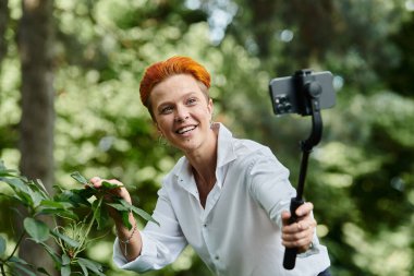 A woman with bright red hair smiles while filming in a lush green outdoor setting. clipart