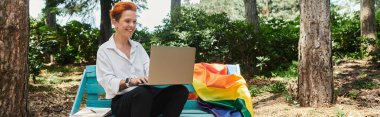 A woman with red hair sits on a park bench working on a laptop, a rainbow flag draped beside her. clipart