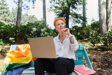 A woman sits on a bench with a laptop, thoughtfully looking up at the trees. clipart