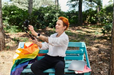 A woman with red hair sits on a bench in a park and takes a selfie with a smartphone. clipart