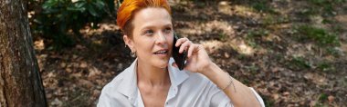 Üniversite kampüsünde telefonla konuşan parlak turuncu saçlı bir öğretmen gülümsüyor..