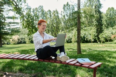 A teacher with red hair smiles while using a laptop on a red wooden bench. clipart