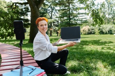 Bir öğretmen kampüs parkında bir bankta oturuyor, elinde bir dizüstü bilgisayarla telefonuyla çekim yapıyor..