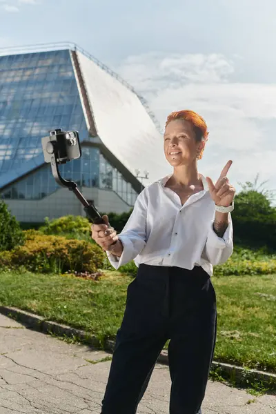 Stock image A woman with short, red hair takes a selfie using a selfie stick in front of a modern building.