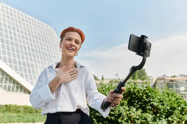 stock image A teacher, dressed in a white shirt and black pants, smiles as she records a selfie video on campus.