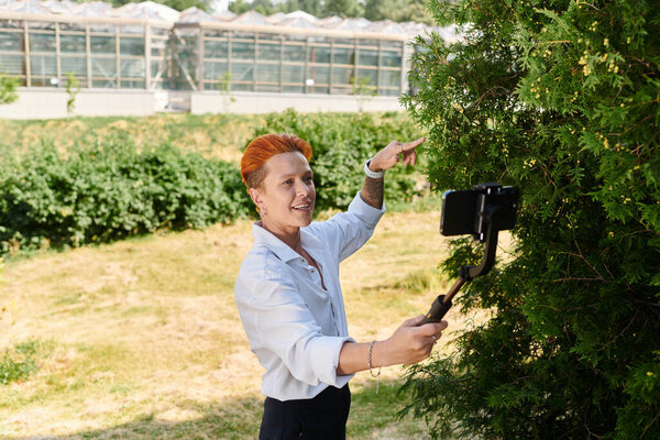 Woman with orange hair films herself on phone by tree on campus