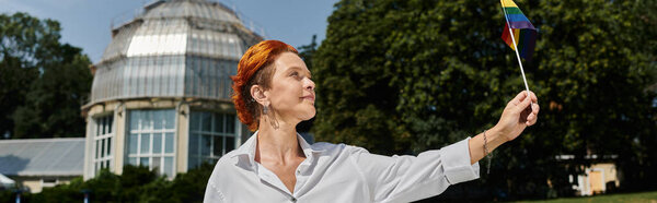 Учитель с рыжими волосами, в белой рубашке, держит радужный флаг на кампусе, улыбаясь.