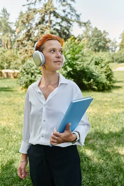 stock image A woman walks on campus, listening to music and holding a blue folder.