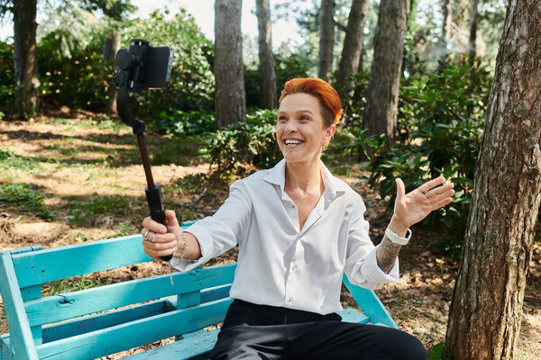 A woman with red hair smiles for a selfie while sitting on a bench in a campus park.