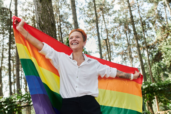 Учительница улыбается, держа в лесу радужный флаг.