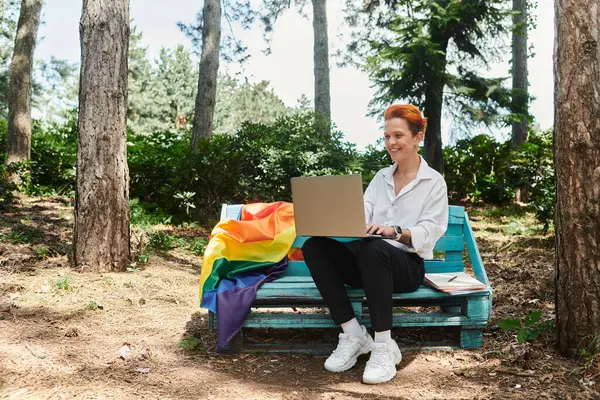 stock image A teacher with red hair sits on a bench in a campus park, using a laptop.