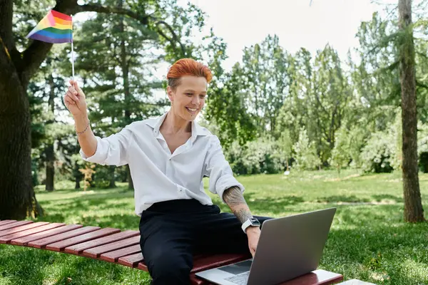 stock image A teacher teacher with a rainbow flag sits on a bench in a park, smiling and working on a laptop.