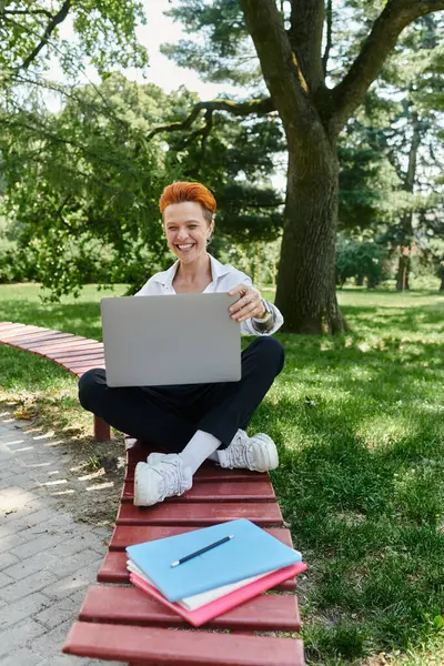 stock image A teacher sits on a campus bench with a laptop, smiling and looking at the screen.