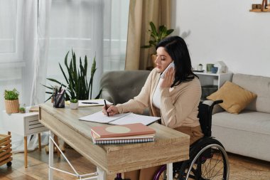 Tekerlekli sandalyedeki bir kadın, işine odaklanmış, masasında otururken telefona cevap veriyor..