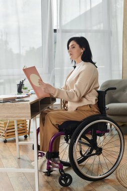 Tekerlekli sandalyede oturan bir kadın pencerenin kenarında oturup kitap okuyor..