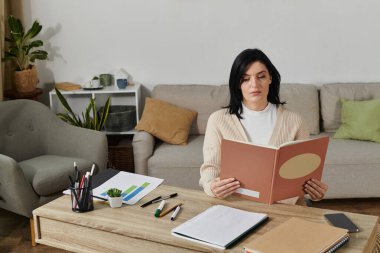 A woman sits at a table in a living room, reading a book. clipart