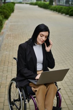 Tekerlekli sandalyedeki bir kadın telefonda konuşurken dizüstü bilgisayarıyla çalışıyor..