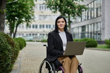 Tekerlekli sandalyedeki bir kadın kucağında dizüstü bilgisayarla kameraya bakıyor..
