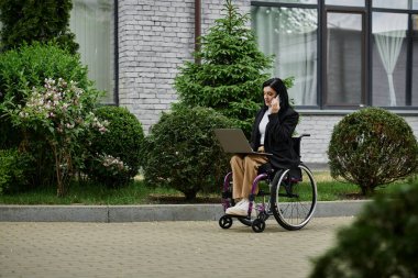 Binanın önünde tekerlekli sandalyede oturan bir kadın dizüstü bilgisayarla çalışıyor ve telefonuyla konuşuyor..