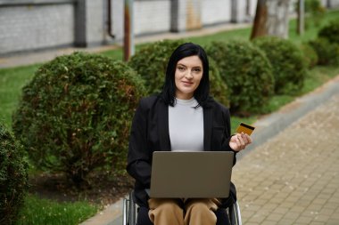 Tekerlekli sandalyedeki bir kadın parkta otururken dizüstü bilgisayar ve kredi kartı kullanıyor..