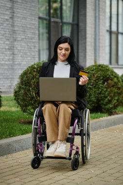 Tekerlekli sandalyedeki bir kadın online alışveriş yapmak için laptop ve kredi kartı kullanıyor..