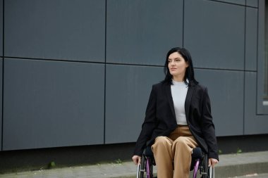Tekerlekli sandalyede takım elbiseli bir kadın şehir ortamında kendinden emin bir şekilde ileriye bakıyor..