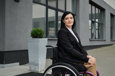 Tekerlekli sandalyedeki bir kadın ofis binasının önünde kendinden emin bir şekilde gülümsüyor..