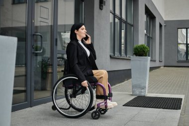 Tekerlekli sandalyedeki bir kadın binanın dışında oturuyor ve telefonuyla konuşuyor..