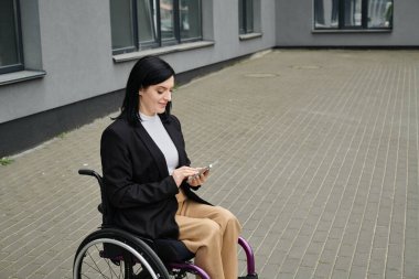 Tekerlekli sandalyedeki bir kadın şehir caddesinde telefonunu kullanırken gülümsüyor..